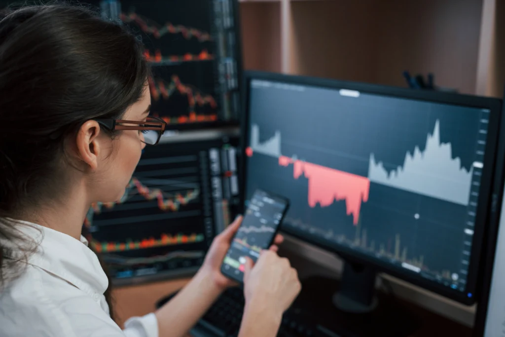 woman-looking-at-index-charts-on-mobile-trading-app-and-monitor-screen – PU Prime | More Than Trading woman looking at indices trading charts on mobile trading app and monitor screen